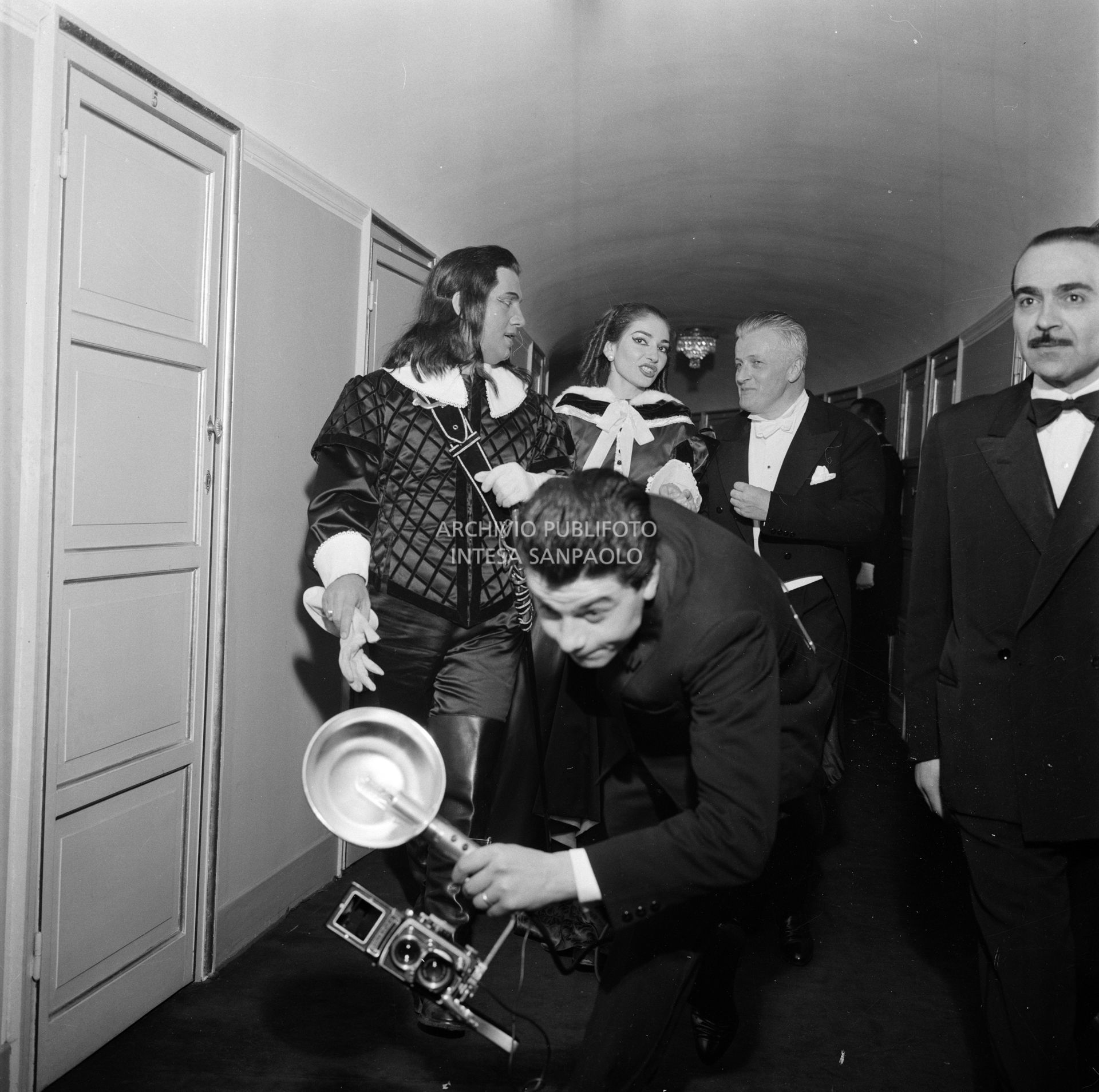 Un fotoreporter con macchina fotografica precede di corsa la soprano Maria Callas, il baritono Ettore Bastianini, in costume di scena, e il direttore d'orchestra Gianandrea Gavazzeni al Teatro alla Scala la  sera dell'inaugurazione della stagione lirica 1957-1958 con l'opera "Un ballo in maschera", di Giuseppe Verdi, diretta dallo stesso Gavazzeni, con la regia di Margherita Wallmann;