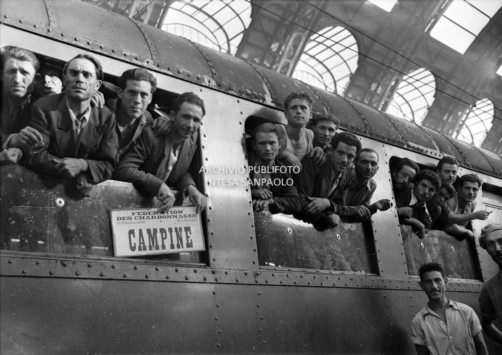 Emigranti, affacciati al vagone ferroviario di un treno, in partenza per le miniere del Belgio, dalla Stazione Centrale di Milano. Sul treno il cartello: "Federation des charbonnages 66, rue des Colonies, Bruxelles. Campine"