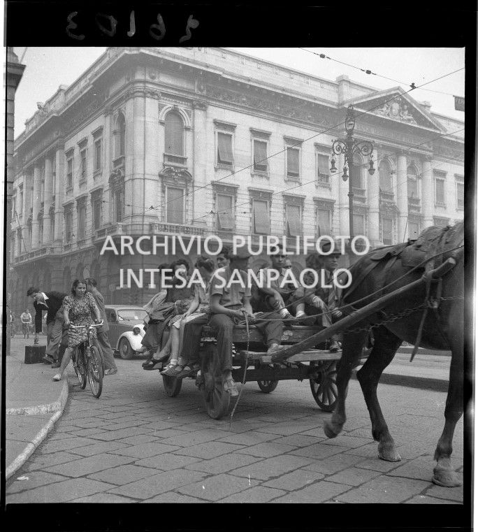 Un carro carico di persone, trainato da un cavallo, in piazza della Scala a Milano. Sullo sfondo il Palazzo della Banca Commerciale Italiana