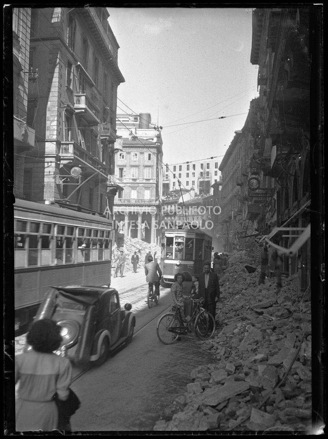 Scorcio di corso Vittorio Emanuele II a Milano verso piazza San Carlo (a sinistra) con il passaggio di tram tipo 1928, civili a piedi, in bicicletta o in automobile; addossate ai margini le macerie degli edifici bombardati