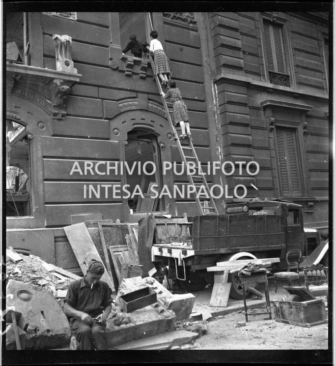 Due donne su una scala, appoggiata su un autocarro, recuperano con l'aiuto di un uomo oggetti da un appartamento in un edificio bombardato e gravemente danneggiato in via Giacomo Leopardi all'angolo con via Aurelio Saffi a Milano