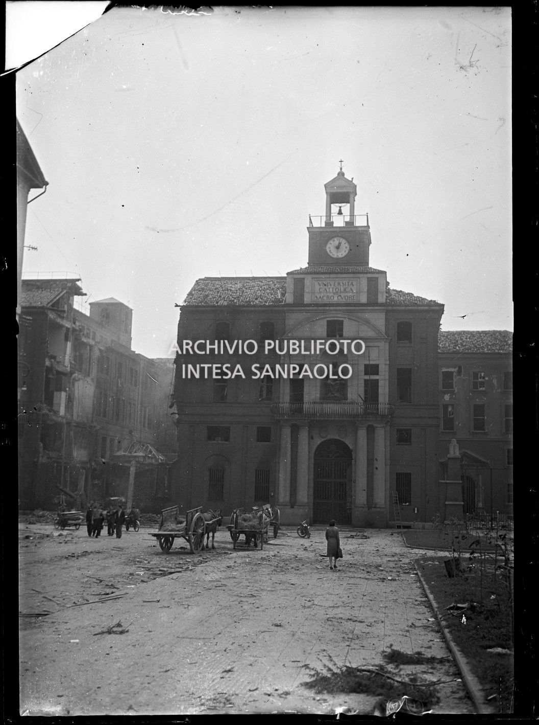 Edificio d'ingresso dell'Università Cattolica del Sacro Cuore, ripreso da piazza Sant'Ambrogio, a Milano, danneggiato dai bombardamenti. Due carri, con cavalli e paglia, sostano nella piazza