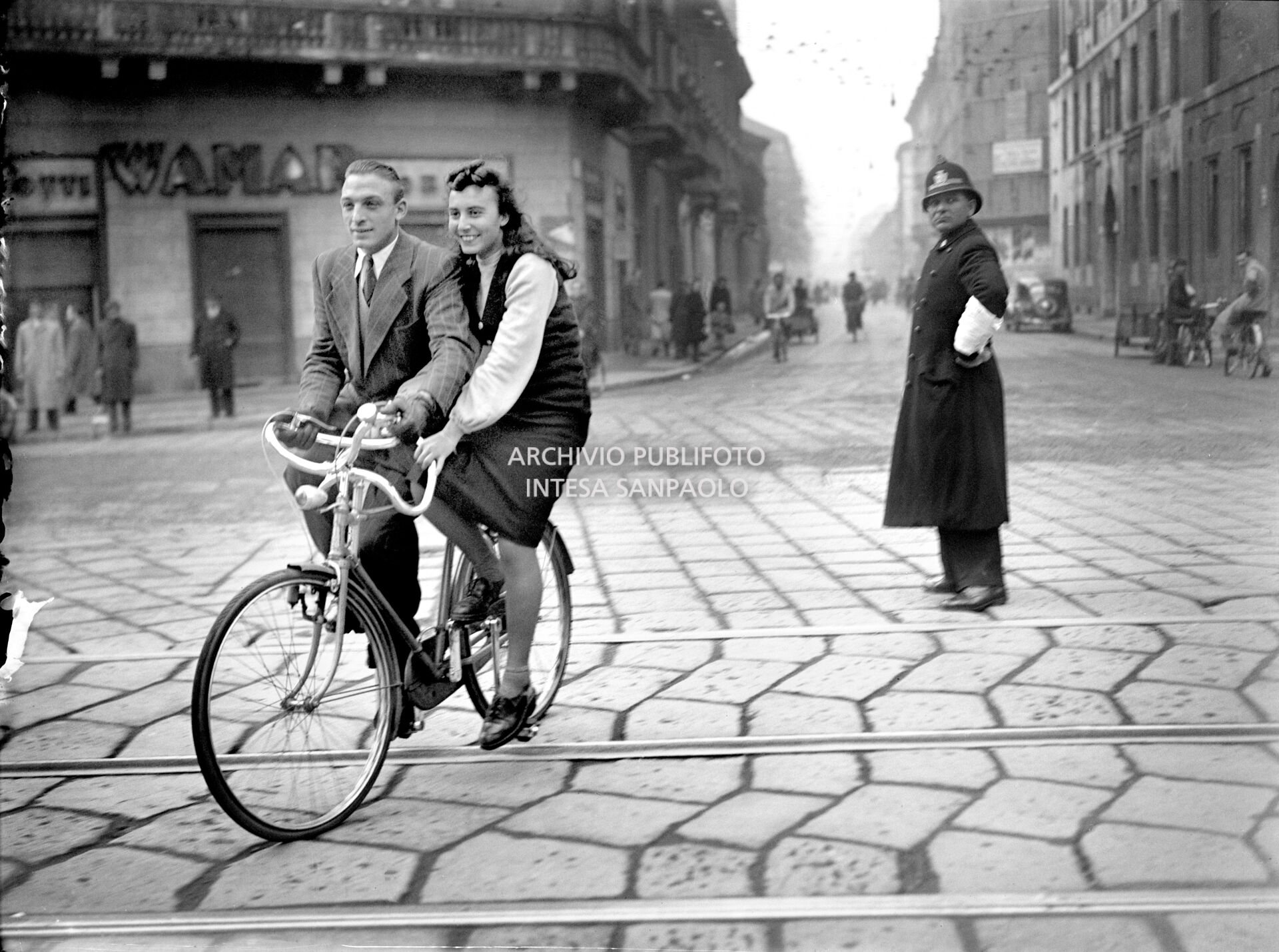 Un uomo e una donna a bordo di un particolare modello di bicicletta in una via di Milano, poco più in là un vigile urbano li guarda incuriosito