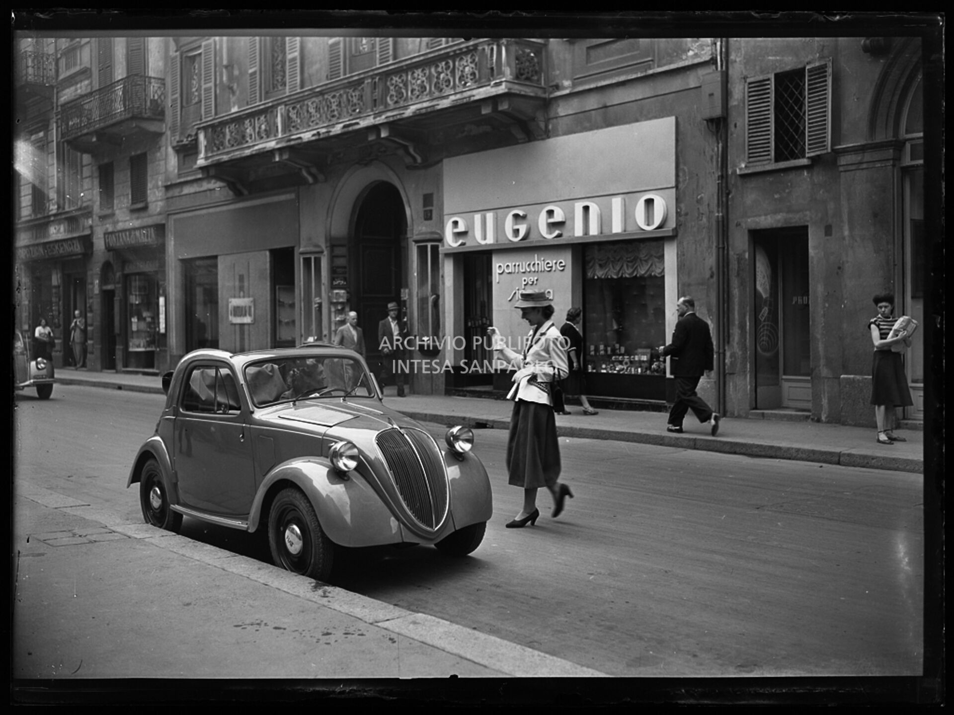 Una donna, in via Monte Napoleone a Milano, si avvicina a una Fiat 500 B. Sullo sfondo il negozio di parrucchiere "Eugenio"