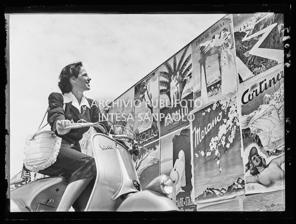 Una donna in Vespa guarda alcuni manifesti di località turistiche