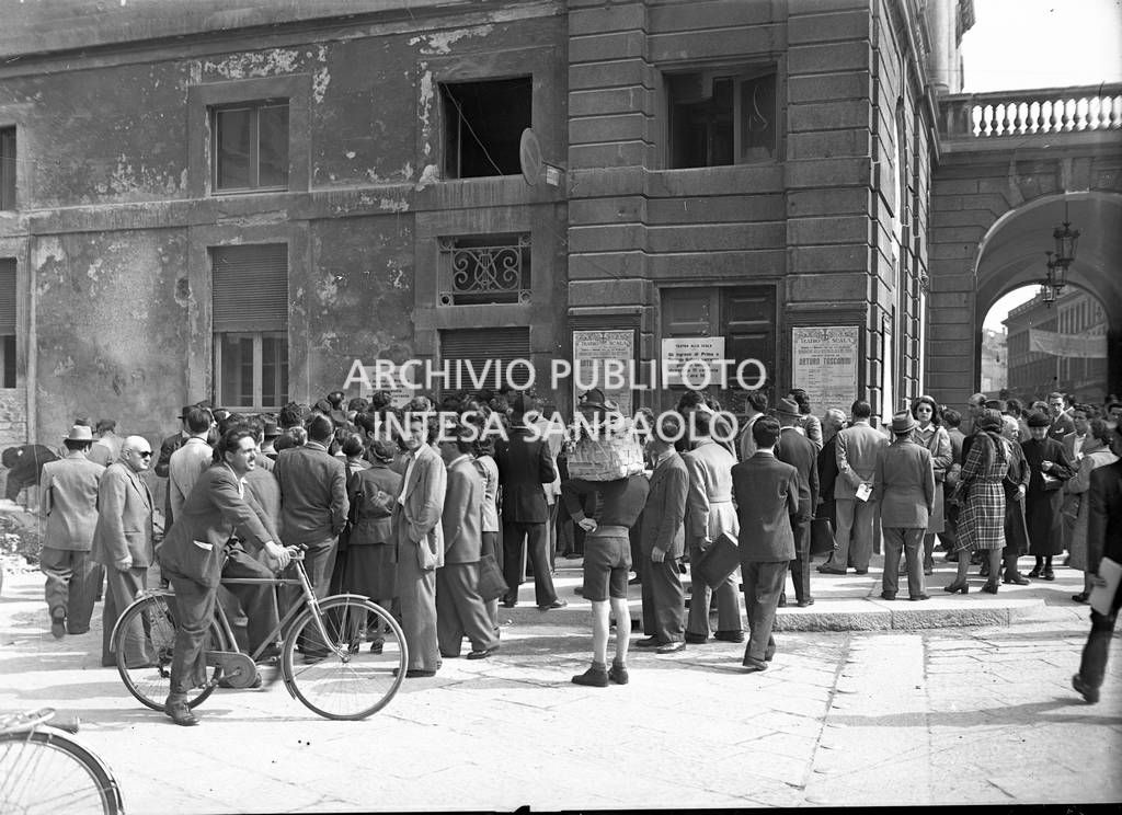 Persone davanti a una delle entrate laterali del Teatro alla Scala su cui sono affissi il manifesto del concerto di riapertura diretto da Arturo Toscanini e alcuni cartelli sulla vendita dei biglietti