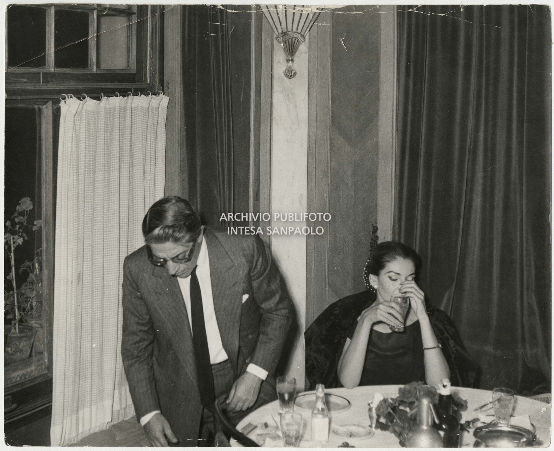Original caption by Publifoto Agency on the back of the print:
						"One of the shots of the "Callas-Onassis affair". The public escapes of the couple that was on everyone's lips began on Saturday 29 August at the "Sea Club" in Monte Carlo. But the first photograph was taken on Thursday 3 September 1959 in Milan. Onassis arrived that afternoon from Venice in his personal plane. The two entered a Milanese night-club [the Montemerlo (Ed.)] at 10 p.m. They dined, danced, drank. They left five hours later and finally smiled at the photographer Nicola Giordano of Publifoto who was waiting for them. A short trip from the night club to the hotel, where the shipowner was staying, for a last "drink". The next day, Friday 4 September, the photographer was still waiting outside the hotel: and at 3.45 p.m. the couple came out and got into the car. Also on Friday. After a stop at the singer's house, here we are at Bresso airport. Maria Meneghini Callas says goodbye to the shipowner who is leaving for Venice in his seaplane. The few people present notice that the lady "is very emotional"."