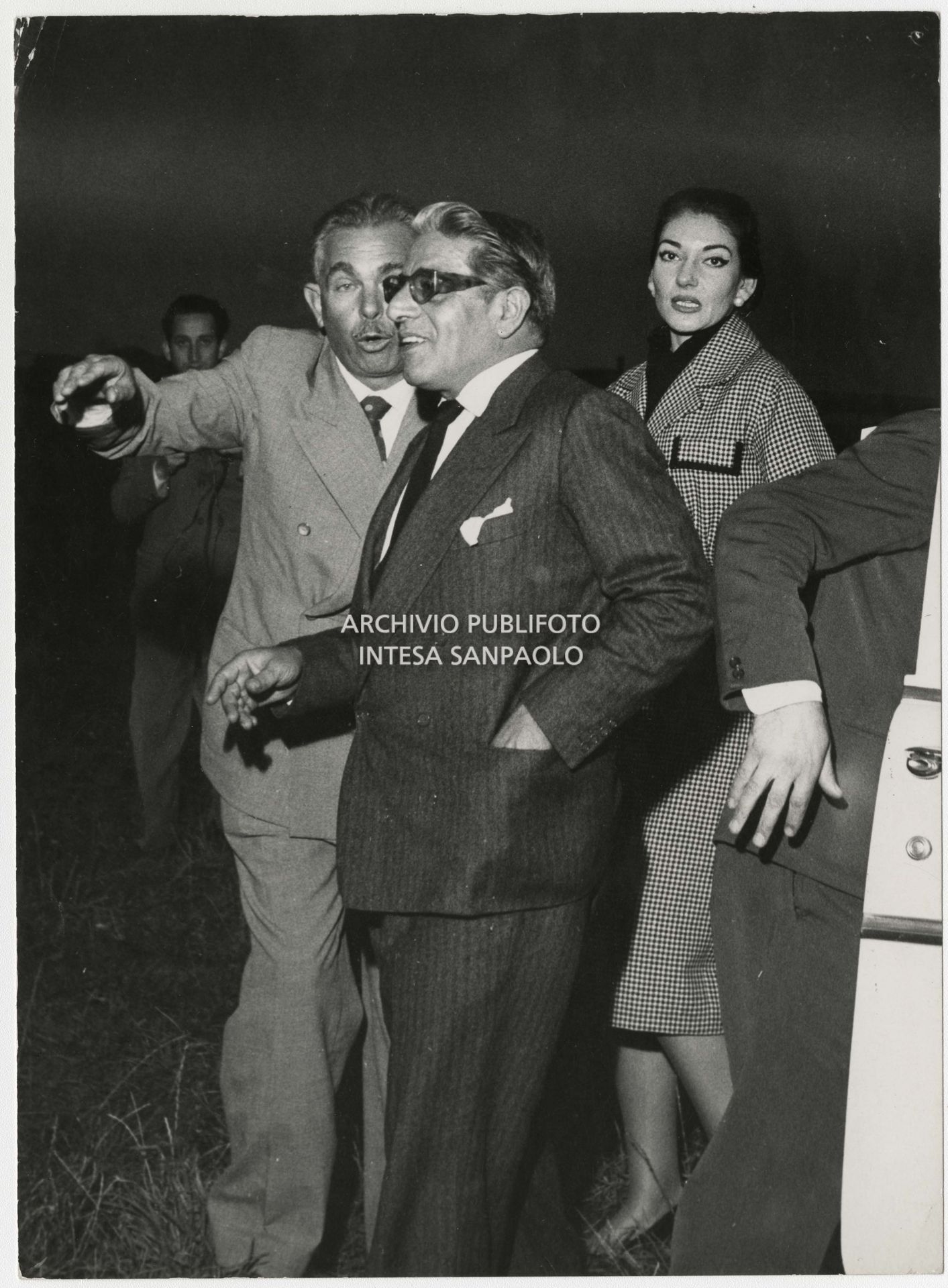 Original caption by Publifoto Agency on the back of the print:
						"One of the shots of the "Callas-Onassis affair". The public escapes of the couple that was on everyone's lips began on Saturday 29 August at the "Sea Club" in Monte Carlo. But the first photograph was taken on Thursday 3 September 1959 in Milan. Onassis arrived that afternoon from Venice in his personal plane. The two entered a Milanese night-club [the Montemerlo (Ed.)] at 10 p.m. They dined, danced, drank. They left five hours later and finally smiled at the photographer Nicola Giordano of Publifoto who was waiting for them. A short trip from the night club to the hotel, where the shipowner was staying, for a last "drink". The next day, Friday 4 September, the photographer was still waiting outside the hotel: and at 3.45 p.m. the couple came out and got into the car. Also on Friday. After a stop at the singer's house, here we are at Bresso airport. Maria Meneghini Callas says goodbye to the shipowner who is leaving for Venice in his seaplane. The few people present notice that the lady "is very emotional"."