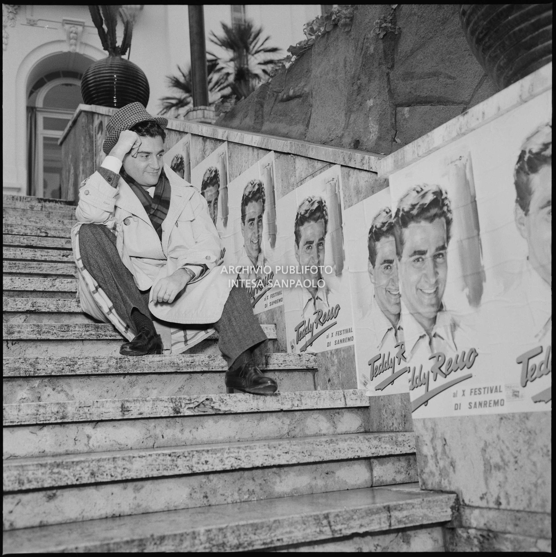 Teddy Reno posing on the Casino steps next to promotional posters featuring him during the 10th Sanremo Festival