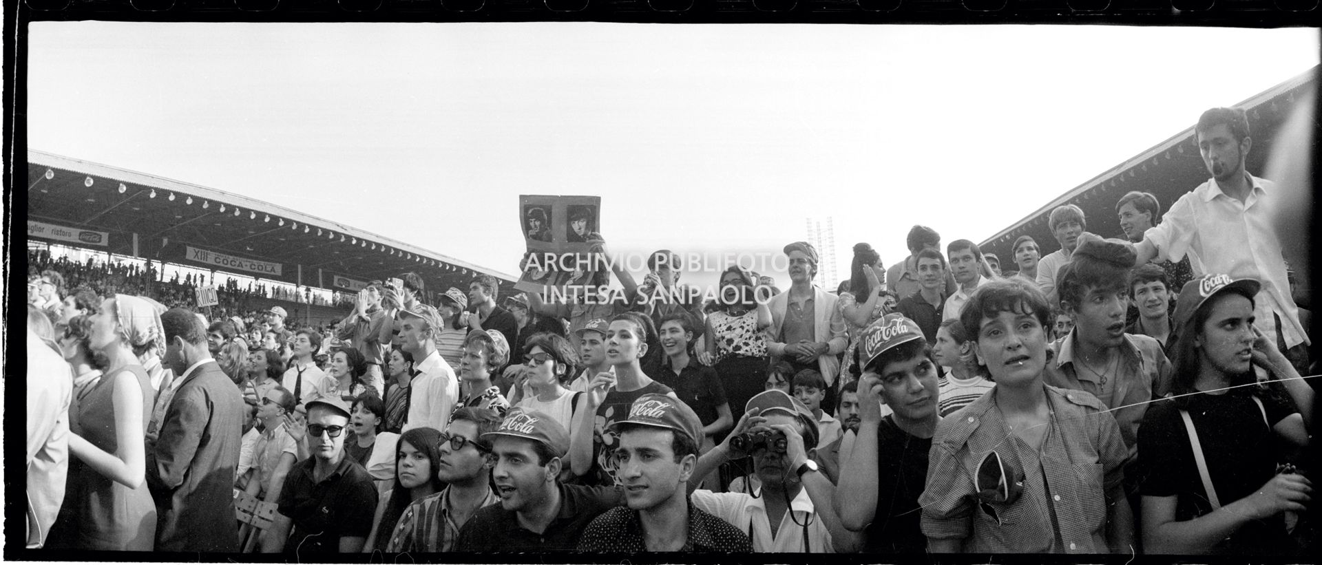Panoramica del pubblico al concerto pomeridiano dei Beatles al Vigorelli di Milano; un ragazzo mostra una fotografia dei musicisti